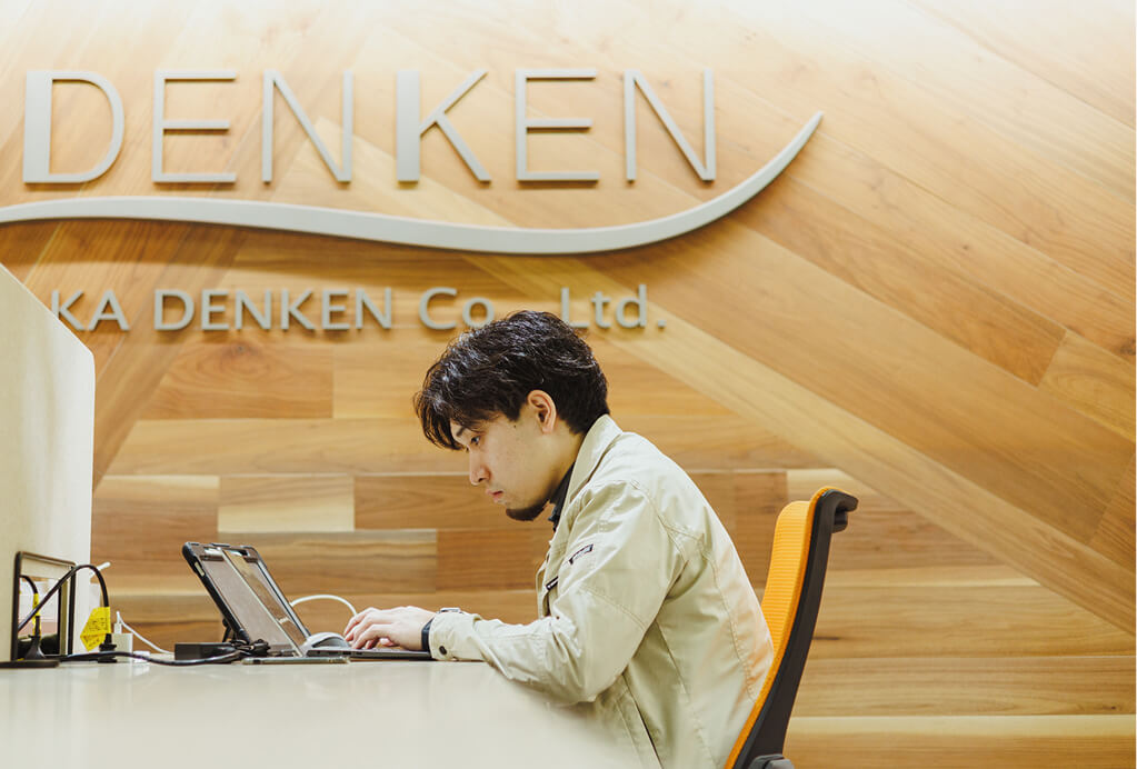 DENKENのサインが入った壁を背にデスクで作業するスタッフ